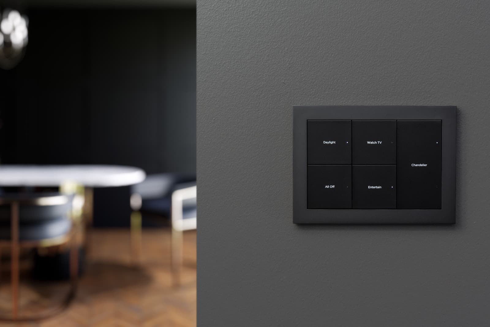 Black scene keypad mounted on a charcoal wall beside a softly focused dining space