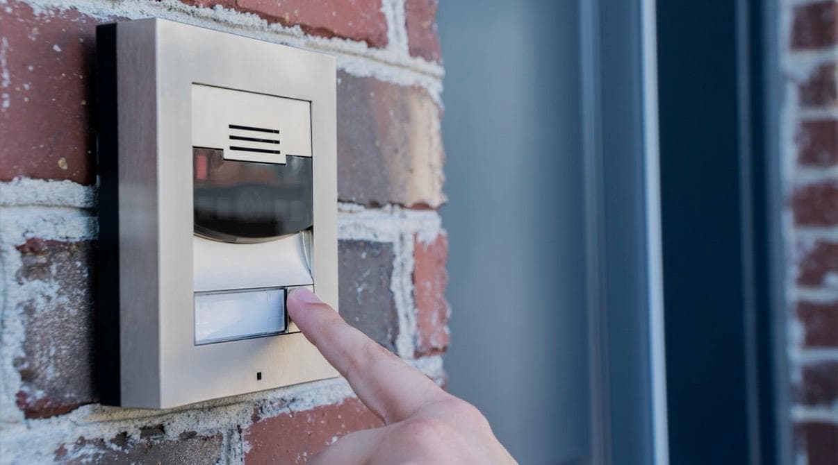 Person pressing a wall-mounted video door station at a brick entrance