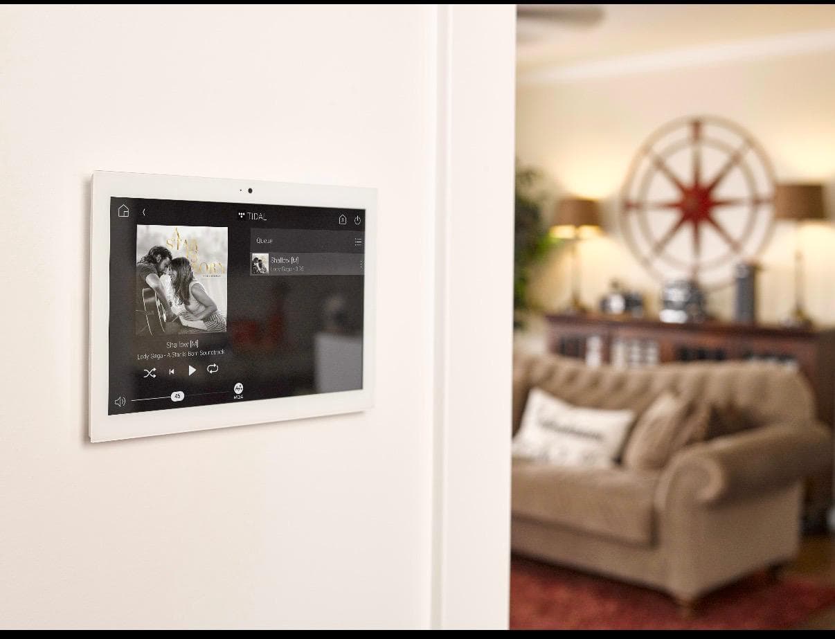 Wall-mounted touchscreen displaying music controls beside a softly furnished living room